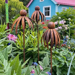 May include: Three rusty metal flower garden stakes with a spherical top and petal-like cutouts. The stakes are set in a lush garden with green foliage and colourful flowers, with a blue house in the background.