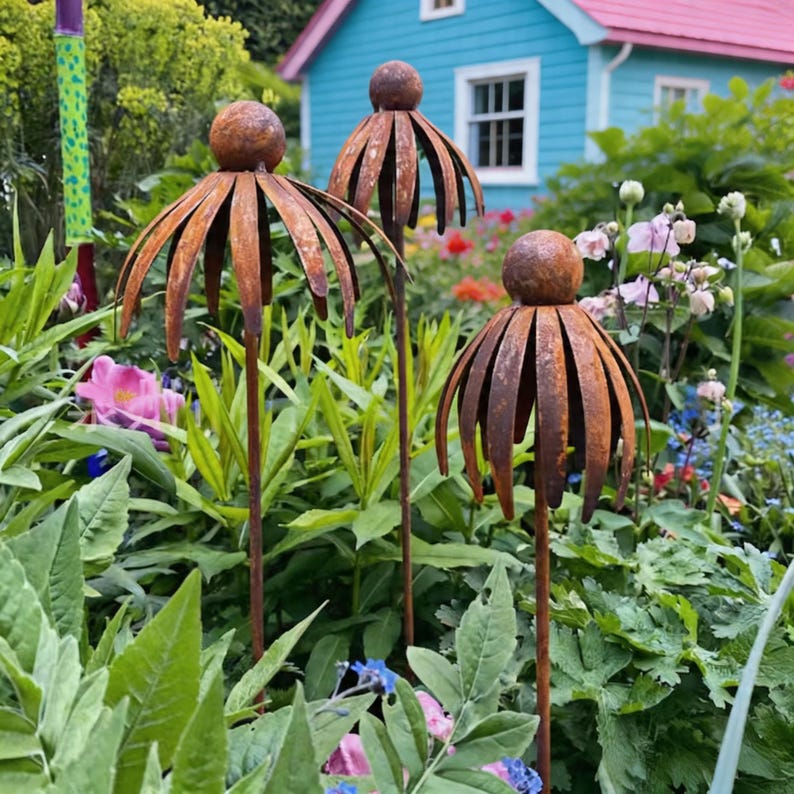 May include: Three rusty metal flower garden stakes with a spherical top and petal-like cutouts. The stakes are set in a lush garden with green foliage and colourful flowers, with a blue house in the background.