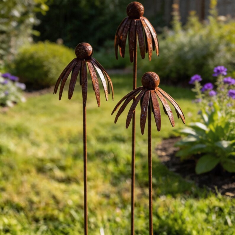 May include: Three rusty metal flower garden stakes with a spherical top and petal-like design. The stakes are placed in a grassy area with a blurred background of greenery and flowers, creating a rustic garden decor.