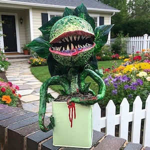 May include: A green, textured, carnivorous plant sculpture with a wide, open mouth featuring sharp, pointed teeth and dark red lips. The plant has large, glossy leaves and is potted in a light green container with red paint drips.