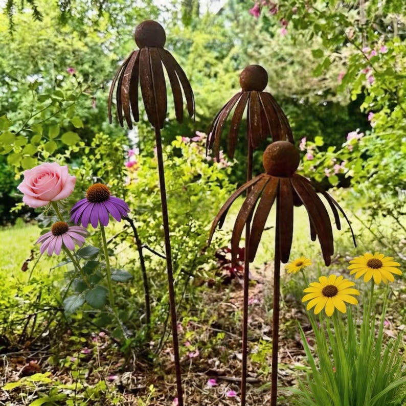 May include: Three weathered metal flower garden stakes with a spherical top and a petal design. The stakes are in a garden with pink and purple flowers and yellow daisies. The background is green foliage.