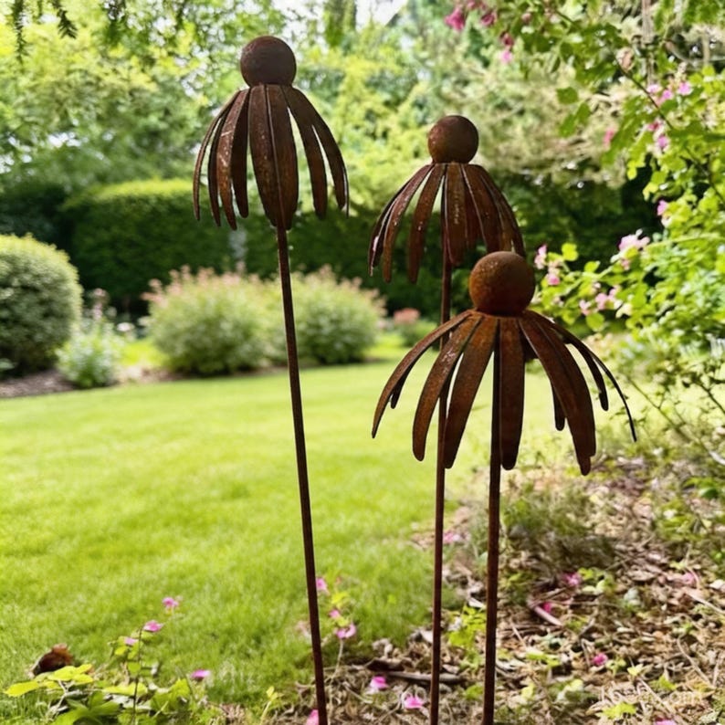 May include: Three rusty metal flower garden stakes with a spherical centre and petal-like edges. The stakes are placed in a grassy yard with green bushes and trees in the background. The flowers are a dark brown colour.