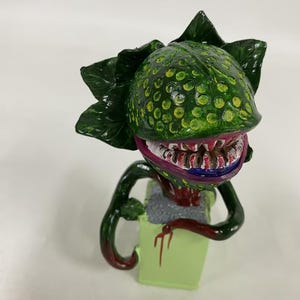 May include: A hand-painted figurine of a carnivorous plant with a large, textured green head featuring yellow spots, sharp white teeth, and a dark red interior. The plant has dark green leaves and a red vine extending from a light green rectangular base.