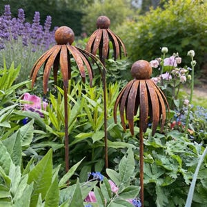 May include: Three weathered metal flower garden stakes with a spherical top and a petal design. The stakes are set in a vibrant garden with green foliage and colourful flowers, including lavender and pink blooms.