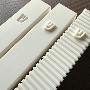 Puede incluir: Tres estuches rectangulares blancos, posiblemente para rollos, con una superficie texturizada y una letra hebrea Shin en relieve. Un estuche tiene un diseño acanalado. Los estuches están dispuestos diagonalmente sobre una superficie de madera oscura.