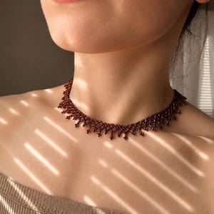 May include: A dark brown beaded choker necklace featuring a delicate, intricate lattice pattern with small, dangling bead accents along the lower edge. The necklace is constructed from numerous tiny, uniform, metallic-finish beads arranged in a repeating geometric design that sits closely around the base of the neck.
