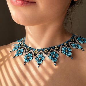 Puede incluir: Un collar tipo gargantilla de cuentas con un patrón geométrico repetitivo en cuentas de semillas turquesas, negras y doradas. El diseño incluye motivos puntiagudos en forma de flor en el borde inferior, acentuados con cuentas transparentes en el borde superior.