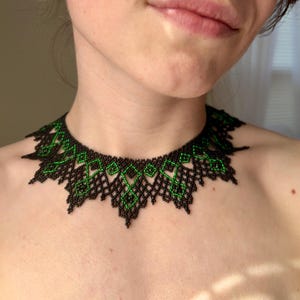 May include: A black and green beaded necklace with a geometric pattern. The necklace has a scalloped edge and is made of small beads. The green beads form a diamond design within the black structure. The necklace is worn around the neck.