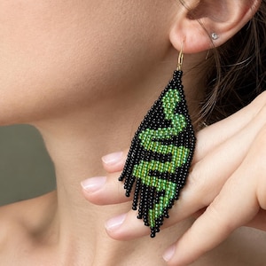 May include: A long, leaf-shaped drop earring crafted from small black beads. The beadwork features a vibrant green snake pattern winding through the centre. The earring hangs from a gold-toned metal hook and includes a fringe of black beads.