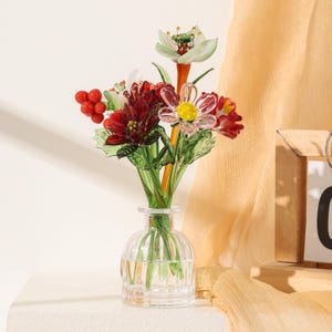 Puede incluir: Un florero de vidrio transparente que contiene un ramo de flores y bayas de vidrio hechas a mano. El arreglo presenta flores de vidrio en tonos rojo oscuro, rosa y blanco con tallos y hojas verdes, todo en un peque&ntilde;o recipiente de vidrio acanalado.