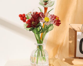 Puede incluir: Un florero de vidrio transparente que contiene un ramo de flores y bayas de vidrio hechas a mano. El arreglo presenta flores de vidrio en tonos rojo oscuro, rosa y blanco con tallos y hojas verdes, todo en un pequeño recipiente de vidrio acanalado.