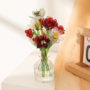 Puede incluir: Un peque&ntilde;o jarr&oacute;n de cristal transparente y acanalado que contiene un ramo de flores de cristal hechas a mano. El arreglo presenta flores de color rojo intenso, una flor blanca con centro amarillo, hojas de cristal verde y bayas rojas.