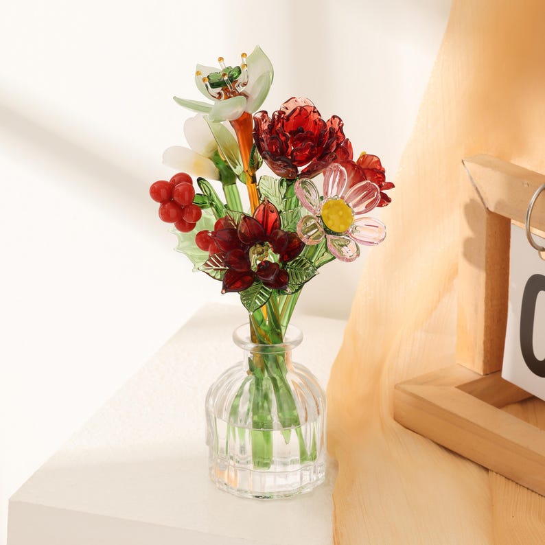 Puede incluir: Un peque&ntilde;o jarr&oacute;n de cristal transparente y acanalado que contiene un ramo de flores de cristal hechas a mano. El arreglo presenta flores de color rojo intenso, una flor blanca con centro amarillo, hojas de cristal verde y bayas rojas.