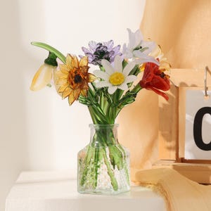 Puede incluir: Un florero de vidrio transparente que contiene un ramo de flores de vidrio hechas a mano, incluyendo una flor amarilla en forma de campana, un girasol dorado, una flor morada, una margarita blanca con centro amarillo, un lirio y una flor roja.