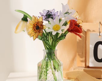 Puede incluir: Un florero de vidrio transparente que contiene un ramo de flores de vidrio hechas a mano, incluyendo una flor amarilla en forma de campana, un girasol dorado, una flor morada, una margarita blanca con centro amarillo, un lirio y una flor roja.
