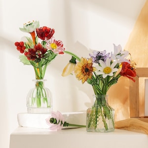 Puede incluir: Dos jarrones de vidrio transparente con arreglos florales de vidrio hechos a mano. El jarr&oacute;n izquierdo contiene tallos florales rojos, blancos y verdes con bayas. El derecho presenta un girasol amarillo, una margarita blanca y p&eacute;talos rojos.