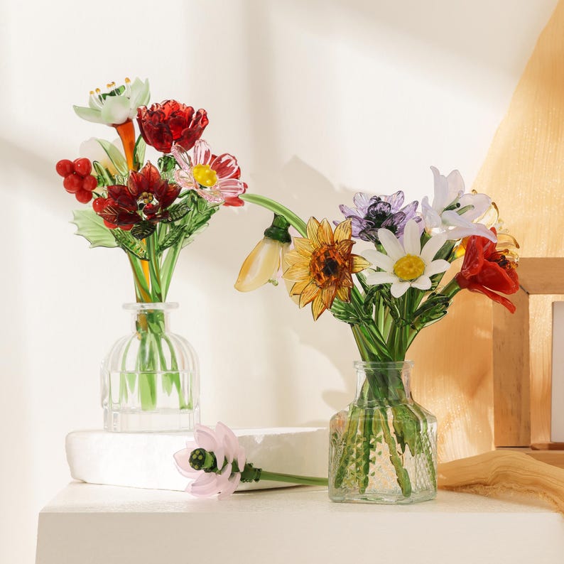 Puede incluir: Dos jarrones de vidrio transparente con arreglos florales de vidrio hechos a mano. El jarr&oacute;n izquierdo contiene tallos florales rojos, blancos y verdes con bayas. El derecho presenta un girasol amarillo, una margarita blanca y p&eacute;talos rojos.