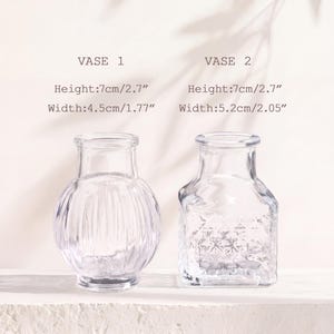 Puede incluir: Dos jarrones de vidrio transparente. El jarr&oacute;n 1 es redondo con estr&iacute;as verticales, 7 cm de alto y 4,5 cm de ancho. El jarr&oacute;n 2 es cuadrado con un patr&oacute;n de diamantes texturizado, 7 cm de alto y 5,2 cm de ancho. Cuellos estrechos y bases anchas.
