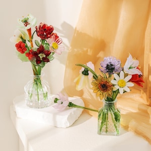 Puede incluir: Dos floreros de vidrio transparente con arreglos florales de vidrio hechos a mano. El florero izquierdo contiene flores y bayas de vidrio rojo, blanco y verde. El derecho presenta un ramo de vidrio amarillo, blanco y morado con una flor rosa.