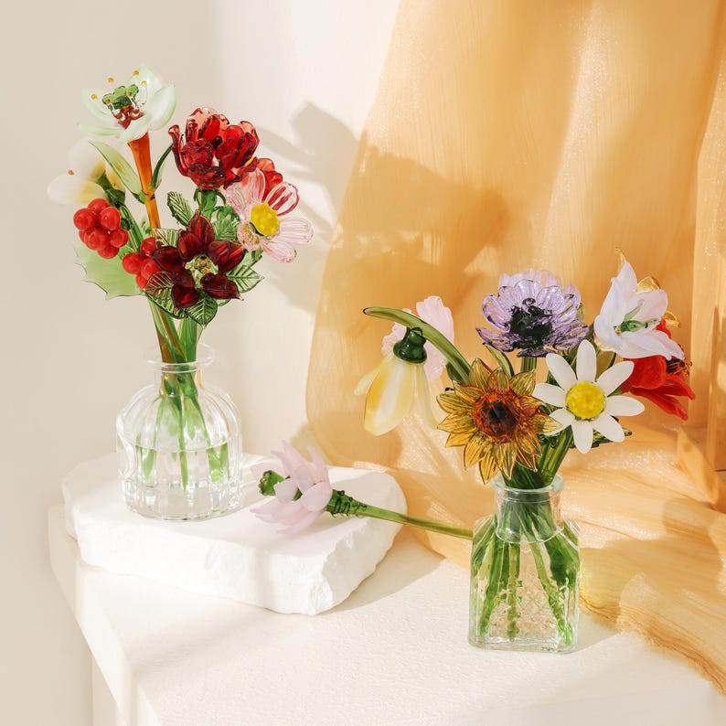 Puede incluir: Dos floreros de vidrio transparente con arreglos florales de vidrio hechos a mano. El florero izquierdo contiene flores y bayas de vidrio rojo, blanco y verde. El derecho presenta un ramo de vidrio amarillo, blanco y morado con una flor rosa.