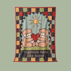Può includere: Una coperta rettangolare con un'illustrazione a cartoni animati di due lontre che tengono un cuore rosso sotto un sole. La coperta ha un bordo a scacchi rosso, nero, verde e giallo, con il testo "Together feels like home" in basso.