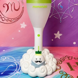 May include: A white and green Mamamoo lightstick featuring a radish-shaped top with a green cap. The handle is white with a small green button. It is mounted on a white, cloud-shaped base featuring a small, cartoonish radish character with a moustache.