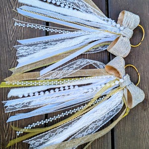 May include: Two decorative wedding tassels with white, gold, and silver ribbons, lace, and pearl accents. Each tassel features a large, sparkly bow and a gold ring for hanging. The tassels are arranged on a wooden surface.