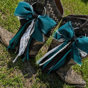 Op de afbeelding: Een paar donkerbruine leren cowboylaarzen met ingewikkelde reliëfpatronen en spitse neuzen. Elke laars is versierd met een grote groenblauwe stoffen strik, witte kanten rand, zwart lint en een snoer van kleine witte kralen aan een zilveren ring.