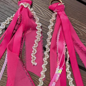 May include: Two gold-toned metal D-rings attached to a collection of hot pink fabric ribbons, frayed chiffon strips, and white decorative trim featuring small faux pearls. The ribbons are layered with iridescent pink sequins and wavy white braid.