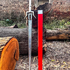 May include: A silver-colored metal sword with a circular pommel and a crossguard featuring a cutout design, accompanied by a long, rectangular red scabbard with a metallic tip. The sword blade is straight and tapers to a sharp point.