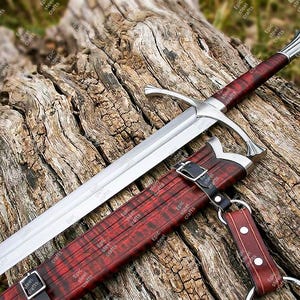 Può includere: Una lunga spada d'argento con un'impugnatura in legno rosso e un fodero abbinato. La spada ha una guardia e una punta. Il fodero è in legno rosso con cinghie in pelle nera. La spada e il fodero poggiano su una superficie di legno stagionato.