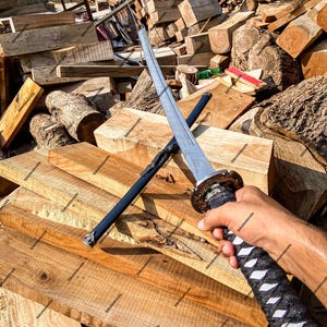 May include: A katana sword with a polished steel blade and a black wrapped handle featuring a decorative metal guard. The sword is positioned diagonally across its black scabbard, which has a silver-toned metal tip and a black cord wrapping.