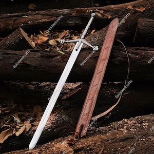 May include: A long, silver sword with a detailed hilt and crossguard rests beside a brown leather sheath. The sword's blade is polished, and the sheath has a decorative pattern. The items are displayed on a bed of dark brown logs.
