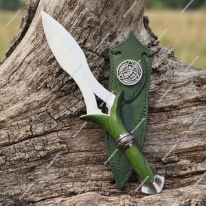 May include: A double-edged dagger with a silver-toned blade and a green handle featuring ornate metal accents. The dagger is paired with a matching green leather sheath that displays a circular silver-toned triquetra emblem.