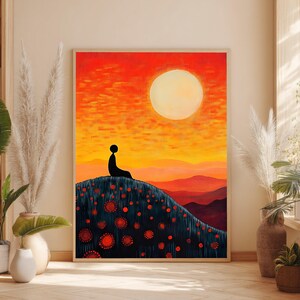 Pode incluir: Uma impressão de arte de parede emoldurada com a silhueta de uma pessoa sentada numa colina escura coberta de flores circulares vermelhas. O fundo exibe um pôr do sol vibrante em laranja e amarelo com um grande sol pálido sobre montanhas distantes.