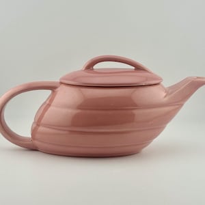 May include: A pink ceramic teapot with a matching lid. The teapot has a curved handle and a spout. The body of the teapot has horizontal ridges. The lid has a small handle for easy removal. The teapot is a solid, muted pink color.