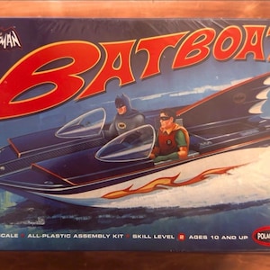 Puede incluir: Una caja de kit de montaje de plástico a escala 1/25 para un modelo de Batboat. La caja presenta una ilustración de Batman y Robin en un barco azul oscuro con calcomanías de llamas naranjas. El texto incluye BATMAN, BATBOAT, SKILL LEVEL 2.