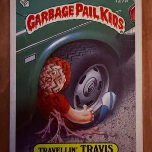 May include: A 1986 Garbage Pail Kids trading card numbered 127a titled Travellin' Travis. The illustration features a character with a tire-tread pattern on their face, positioned next to a green car tire. The card includes the text Peel Here.