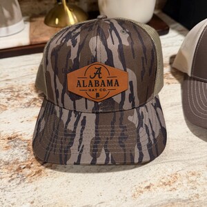 May include: Camouflage-patterned baseball cap with a brown leather patch featuring the text "ALABAMA HAT CO." The cap has a curved brim and a mesh back. The hat is designed for outdoor activities and casual wear.