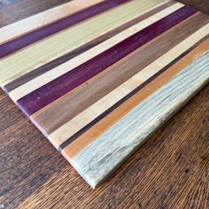 May include: A rectangular chopping board with a striped design. The board features alternating stripes of various wood tones, including light beige, purple, brown, and light blue. The wood grain is visible, adding texture to the surface.