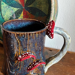 May include: A ceramic mug with a dark blue and brown wood grain texture. The exterior features three-dimensional red mushrooms with white spots attached to the side and the handle. The interior of the mug is a deep, glossy dark blue glaze.