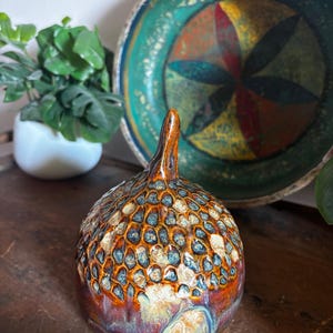 May include: A ceramic acorn-shaped sculpture with a glossy, multi-colored glaze featuring shades of brown, blue, and cream. The top section has a textured, honeycomb-like pattern, while the base is smooth. The text "Large Woodland" is visible.