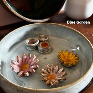 May include: A light blue ceramic bowl containing two small round vessels and three flower-shaped ceramic pieces in pink, yellow, and white with yellow centers. The text Blue Garden is printed in white on the right side of the image.