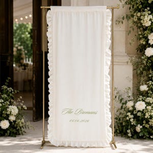 May include: Cream-colored wedding welcome sign with ruffled edges, displayed on a gold metal stand. The sign reads "The Bromans" and "08.06.2026" in green script. The scene is set outdoors with floral arrangements.