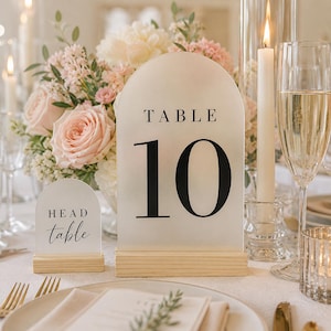 May include: Frosted acrylic table number 10 with a wooden base. The number is in a bold black font. A smaller sign reads "Head Table" in script. The signs are on a table set for a wedding, with flowers and candles.