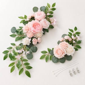Op de afbeelding: Twee bloemstukken met roze en witte rozen, pioenrozen en groen. De arrangementen bevatten eucalyptusbladeren en kleine roze bloemen. Ritssluitingen en clips zijn ook inbegrepen.