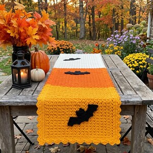 Peut inclure: Un chemin de table rectangulaire en crochet avec un design en blocs de couleurs blanc, orange et jaune. Le chemin est décoré de trois silhouettes de chauves-souris en feutre noir placées au centre. Il présente une bordure festonnée.