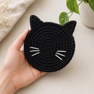 May include: A circular black crochet coaster shaped like a cat head with two pointed ears at the top. The coaster features three white embroidered stitches on each side to represent whiskers, creating a simple and functional feline-inspired design.