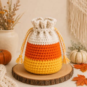 Puede incluir: Una bolsa de ganchillo con cordón que presenta un patrón de colores de maíz dulce con una sección superior blanca, una sección central naranja y una sección inferior amarilla. La bolsa incluye dos cordones trenzados en blanco y amarillo.