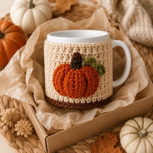 May include: A white ceramic mug featuring a beige crocheted cozy with a three-dimensional orange pumpkin applique. The pumpkin has a dark brown stem and a small green vine detail. The mug is nestled inside a brown cardboard box with crinkled paper.